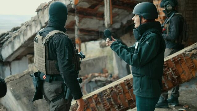A female war presenter in protective body vest and a helmet interviews with a soldier at the scene of an attack in the ruins of a building in a war zone.