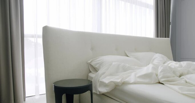 Detail of interior in modern bedroom, unmade bed with upholstered headboard and bedding linen, night side table and window with tulle and curtains. Comfortable bed with pillows and soft duvet