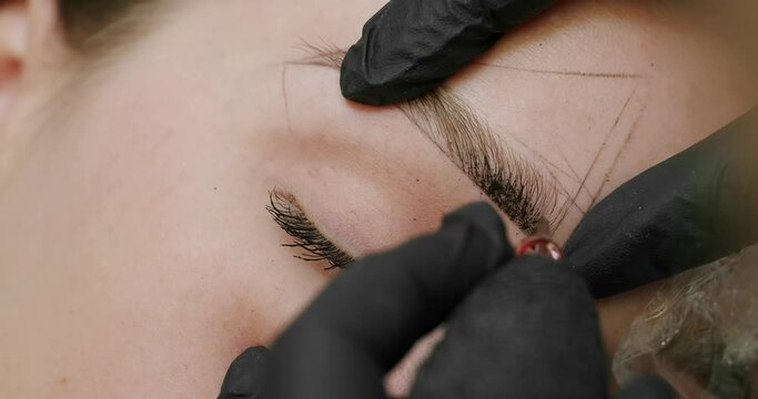 Closeup on woman client face, beauty master doing permanent makeup of eyebrows. Young female on brows microblading procedure at salon. cosmetician using professional apparatus for brows tattooing