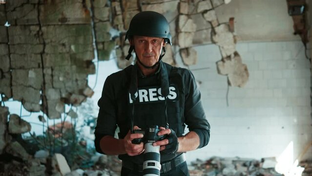 Portrait of a mature man in helmet and bulletproof vest taking photos in war zone, holding digital camera and looking at camera 
