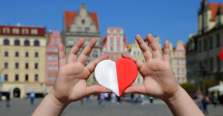 A red-white heart in the colors of the national flag of Poland in the hands of a child against the backdrop of the old town square. Independence Day of Poland. Freedom and Democracy