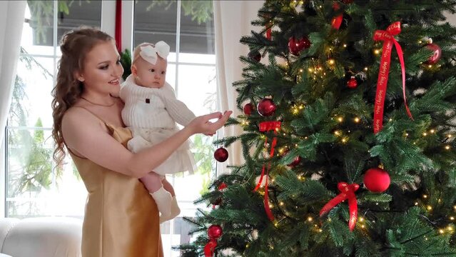 A beautiful mother with a little daughter in her arms decorates the Christmas tree in the living room. New Year's gifts under the tree. large bright room prepared for Christmas. sweet family. High qua
