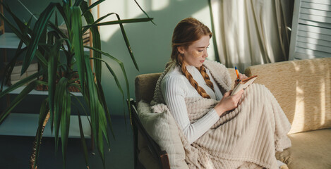 Red haired woman studying from home in comfortable warm white winter blanket, writing ideas on a...