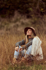 A smiling girl, keeping her eyes closed, dressed casually, wearing a hat, sitting in the field.