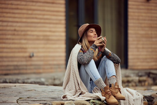 A happy girl, drinking a coffee, sitting in front of the cottage, covered in a blanket.