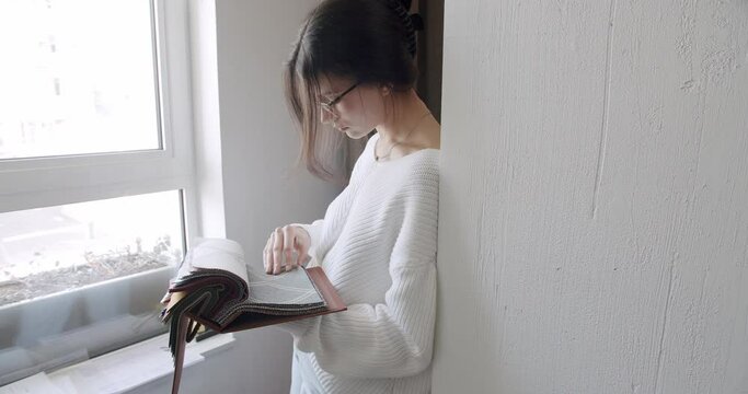 Young woman in eyeglasses hold catalog with fabric samples and choosing color for curtains. Female designer select textile swatch for home decoration. Renovation of apartment concept