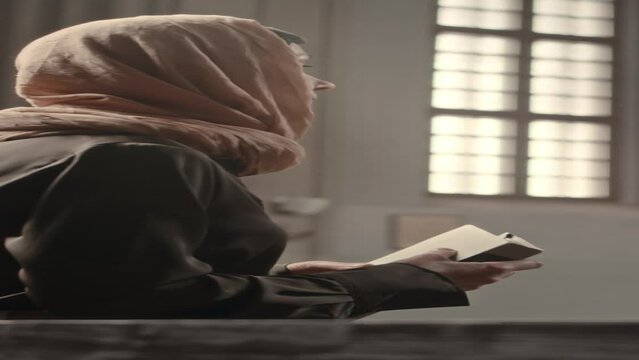 Vertical shot of mature Caucasian female parishioner in headscarf reading Bible in Catholic Church
