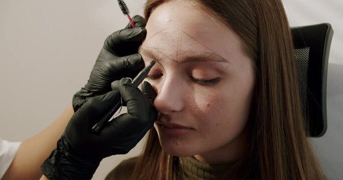 Beauty master doing shape of eyebrows for client woman before henna dyeing. Closeup on female face with prepared brow contour for microblading or permanent makeup at salon