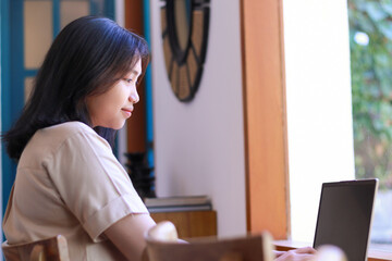 happy asian woman working on laptop sitting near window of coffee shop wearing casual clothes,...