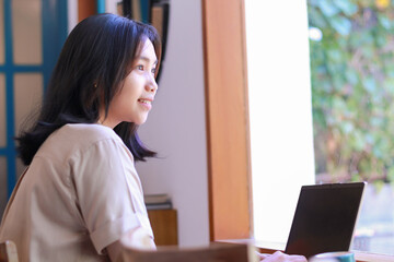 happy asian woman working on laptop sitting near window of coffee shop wearing casual clothes, female designer have online meeting project in cafe