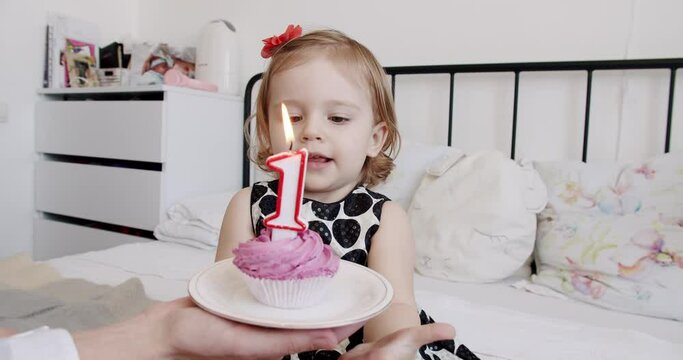Cute baby girl in dress celebrating birthday, blowing out candle on cupcake and excited clapping hands while sitting on bed in kids room. Happy toddler celebrate one year with family at home.