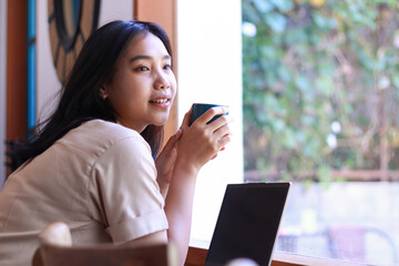 happy asian woman sitting in cafe drinkking coffee and working with laptop near window wearing fashionable clothes, contemplative female remote worker holding mug in coffee shop look away