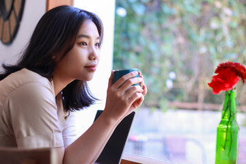 happy asian woman sitting in cafe drinkking coffee and working with laptop near window wearing fashionable clothes, smiling female remote worker holding mug in coffee shop look away