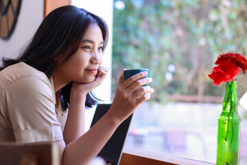 happy asian woman sitting in cafe drinkking coffee and working with laptop near window wearing...