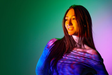 Beautiful young woman posing against gradient pink green background with neon reflection on face...