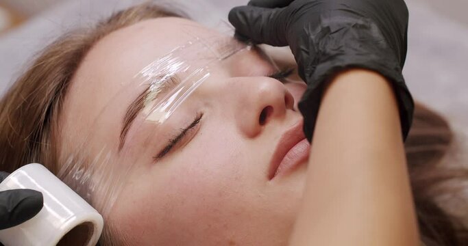 Beautician making permanent makeup to woman client, applying analgesic and cover eyebrow with film. Closeup on female face during brows tattooing at beauty salon.