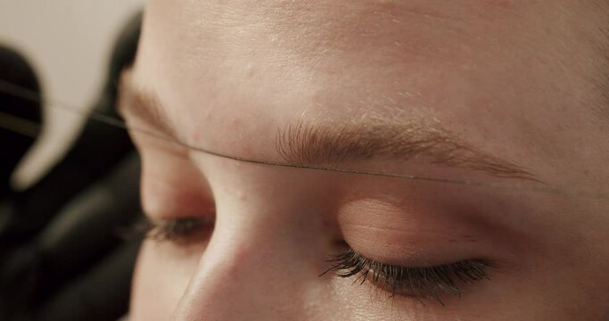 Beautician in gloves using thread to measurements correct shape of eyebrows before microblading procedure in beauty salon. Closeup on young female client face on permanent make-up.