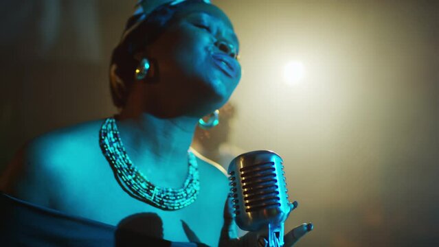 Chest up of artistic young Black female jazz vocalist wearing vintage beads necklace and patterned headscarf singing in retro microphone in blue light of projector