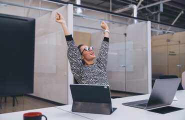 Excited woman raising arms up while sitting at table with laptop and tablet