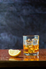 A glass of whiskey with lemon and ice stands on old polished table ion dark background