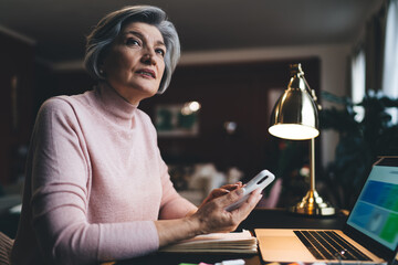 Thoughtful senior woman with smartphone sitting at table