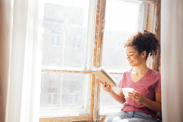 Young smiling pretty mixed race girl sits on windowsill with porcelain cup of tea and book