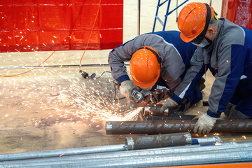 Two workers are sawing pipe. Craftsmen produce steel pipeline. Process of installing pipes inside building. Construction of engineering communications. Factory craftsmen sawing off piece of pipe - obrazy, fototapety, plakaty