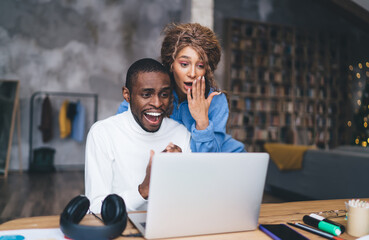 Joyful Black man in white turtleneck and woman in blue top share excitement over laptop screen