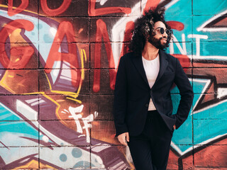 Handsome hipster model. Arabian man dressed in suit jacket clothes. Fashion male with long curly hairstyle posing in street near graffiti wall