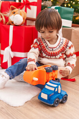 Adorable toddler playing with truck toy sitting on floor by christmas tree at home