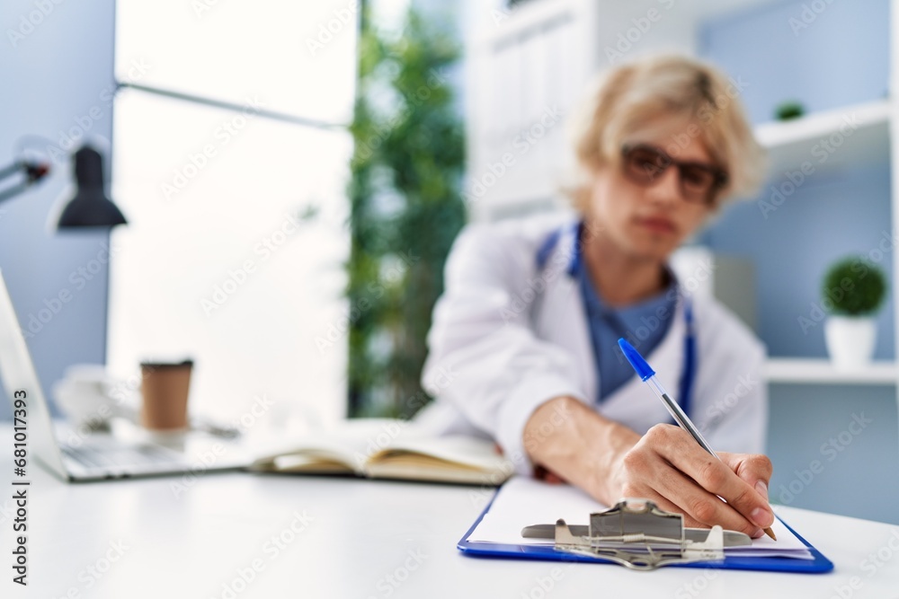 Poster young blond man doctor using laptop writing on document at clinic