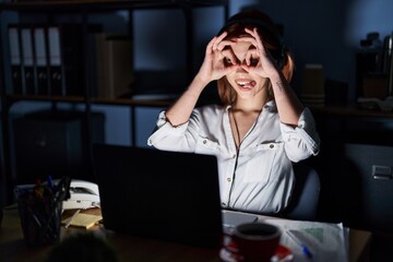 Young caucasian woman working at the office at night doing ok gesture like binoculars sticking tongue out, eyes looking through fingers. crazy expression.