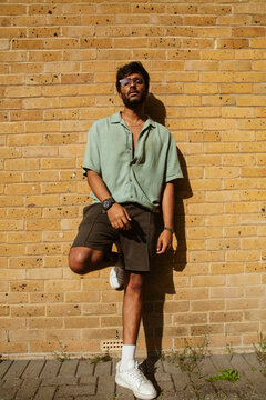 Young Indian man in fashionable outfit leaning against brick wall and looking into camera