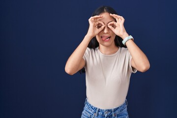 Young hispanic woman standing over blue background doing ok gesture like binoculars sticking tongue out, eyes looking through fingers. crazy expression.