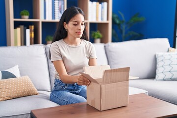 Young hispanic woman opening cardboard box relaxed with serious expression on face. simple and natural looking at the camera.