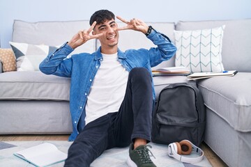 Young hispanic man sitting on the floor studying for university doing peace symbol with fingers...
