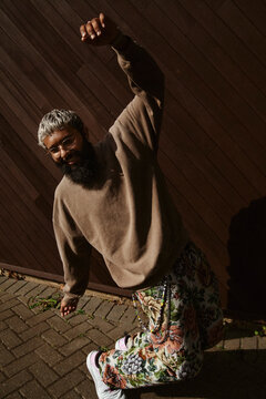 Happy young Tamil guy in trendy outfit dancing in front of brown wall