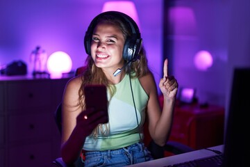 Young hispanic woman playing video games with smartphone smiling with an idea or question pointing...