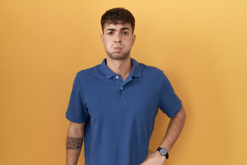Young hispanic man standing over yellow background puffing cheeks with funny face. mouth inflated with air, crazy expression. - obrazy, fototapety, plakaty