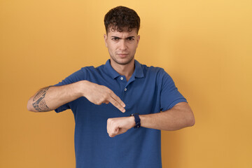 Young hispanic man standing over yellow background in hurry pointing to watch time, impatience, upset and angry for deadline delay