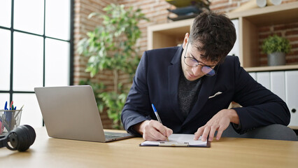 Fototapeta premium Young hispanic man business worker using laptop writing on clipboard at office