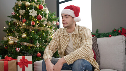 Obraz na płótnie Canvas Young hispanic man sitting on sofa by christmas tree looking upset at home