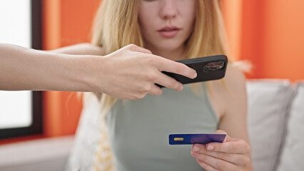 Young blonde woman make photo to credit card by smartphone sitting on sofa at home