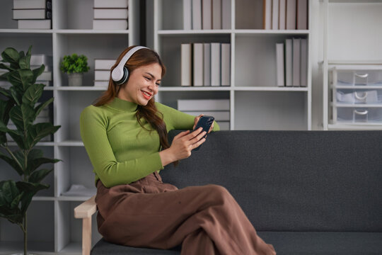Happy Asian woman listening to music on mobile phone while sitting on sofa at home Relaxed smiling girl with headphones in the morning It's time to rest.