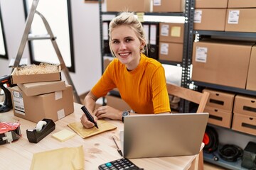 Young blonde woman ecommerce business worker using laptop writing on package at office
