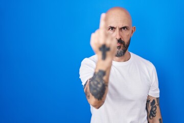 Hispanic man with tattoos standing over blue background showing middle finger, impolite and rude...