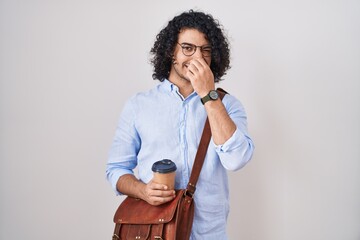 Hispanic man with curly hair drinking a cup of take away coffee smelling something stinky and disgusting, intolerable smell, holding breath with fingers on nose. bad smell - obrazy, fototapety, plakaty
