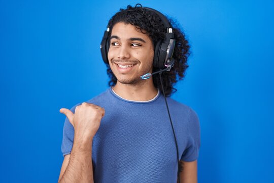 Hispanic man with curly hair listening to music using headphones smiling with happy face looking and pointing to the side with thumb up.