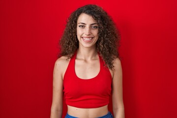 Hispanic woman with curly hair standing over red background with a happy and cool smile on face....