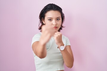 Hispanic young woman standing over pink background ready to fight with fist defense gesture, angry and upset face, afraid of problem